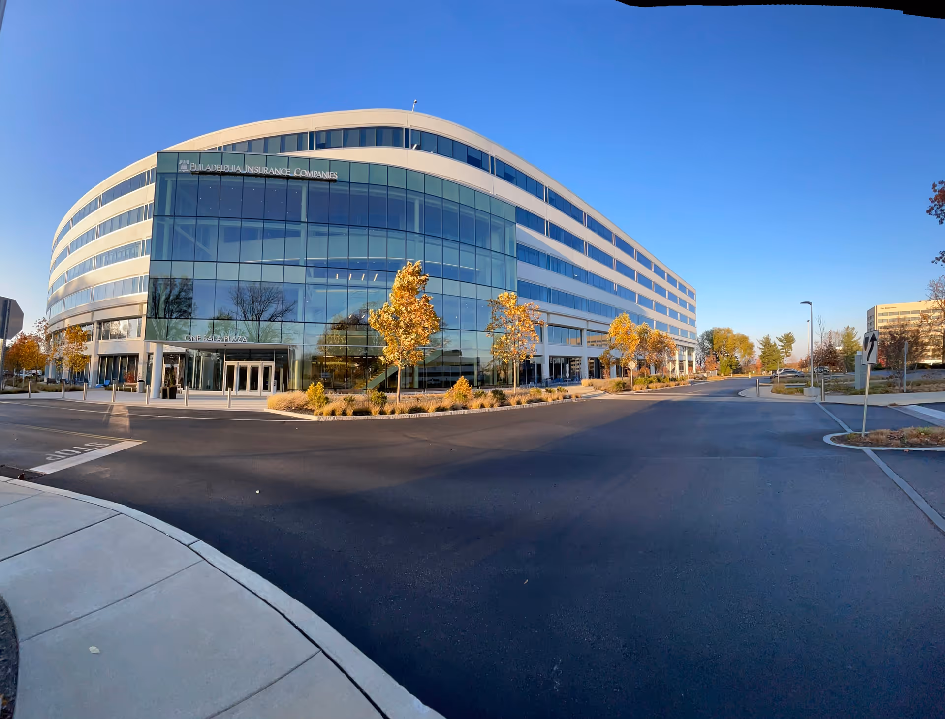 Curved glass-front building with a broad street and autumn trees.