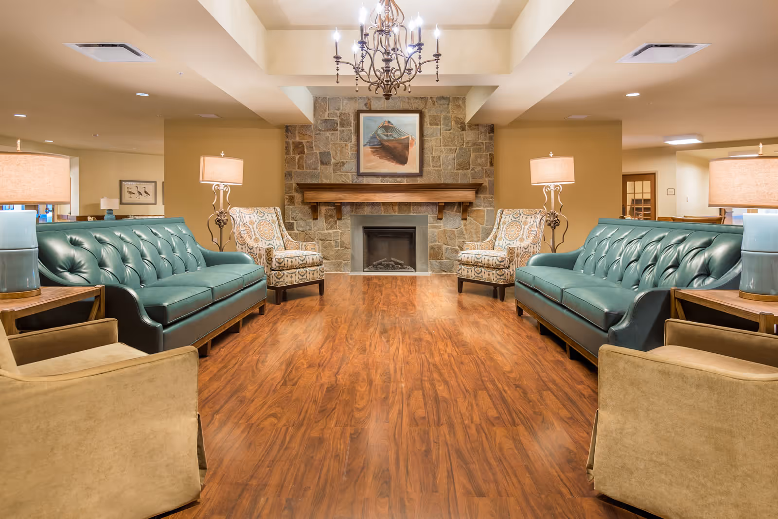 A cozy senior living community lounge featuring two teal leather sofas facing each other, two patterned armchairs, and two beige armchairs. The room has a wooden floor, a stone fireplace with a wooden mantle, and a painting of a boat above the fireplace. A chandelier hangs from the ceiling, and table lamps with beige shades are placed on wooden side tables beside the sofas.