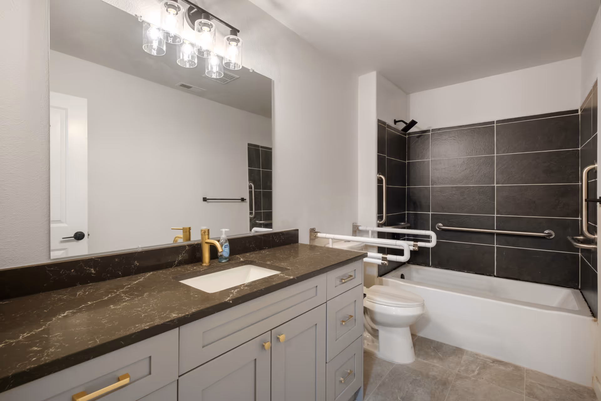 Modern accessible bathroom with a single sink vanity and marble countertop, large mirror, and a bathtub with dark tile surround and multiple grab bars.
