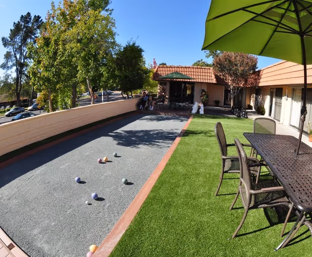 Outdoor courtyard at a care center featuring a bocce ball court, artificial turf, a patio dining set with umbrella, and a single-story building with a red tile roof.
