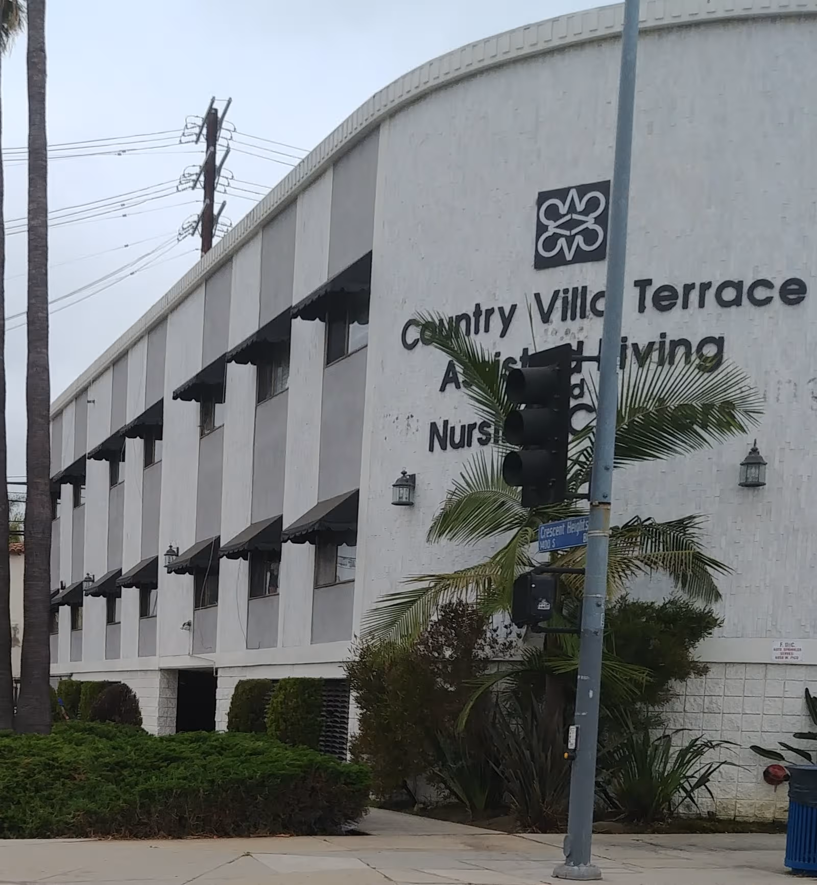 Exterior view of a white assisted living building with awnings, palm trees, and a traffic light.