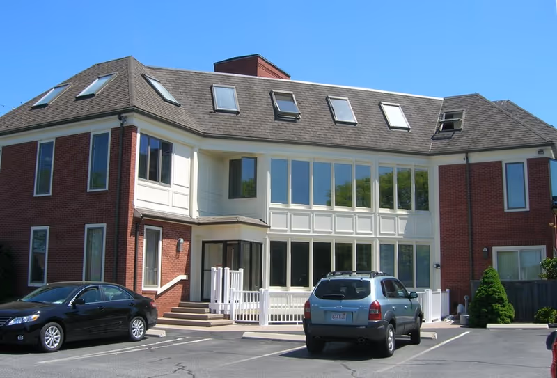 Two-story brick senior living facility with large windows and parked cars in a lot.