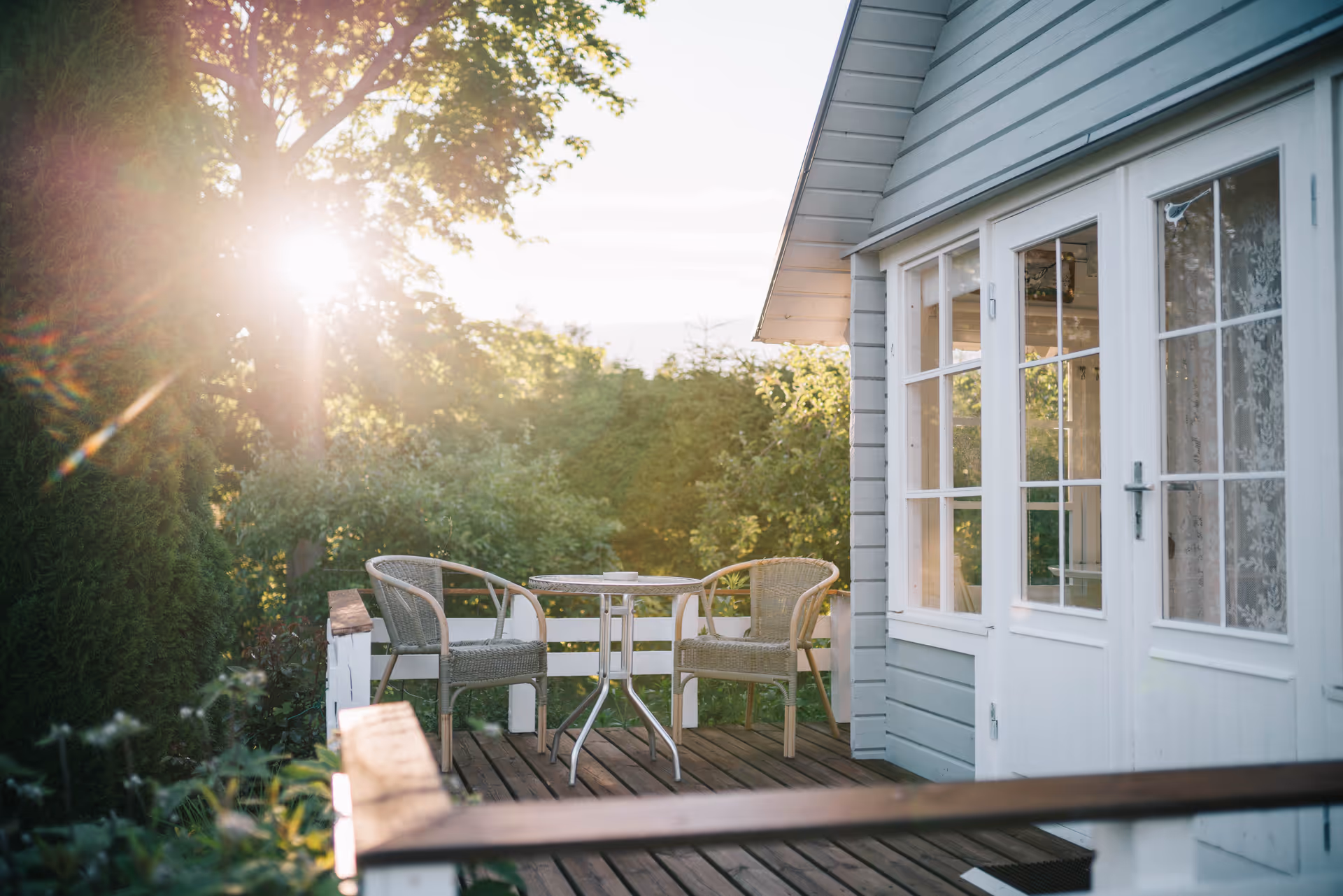 Sunlit outdoor deck with a small table and wicker chairs beside a light blue building.