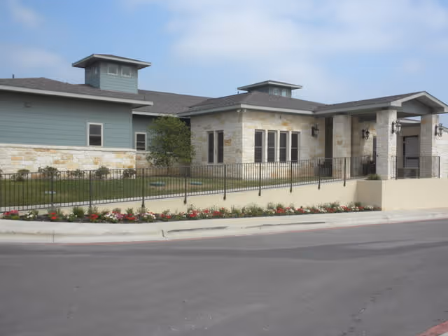 Exterior view of a single-story building with light stone and blue siding, featuring multiple windows and two small tower-like structures on the roof. The building is surrounded by a black metal fence and a landscaped area with grass and flowers along the sidewalk.