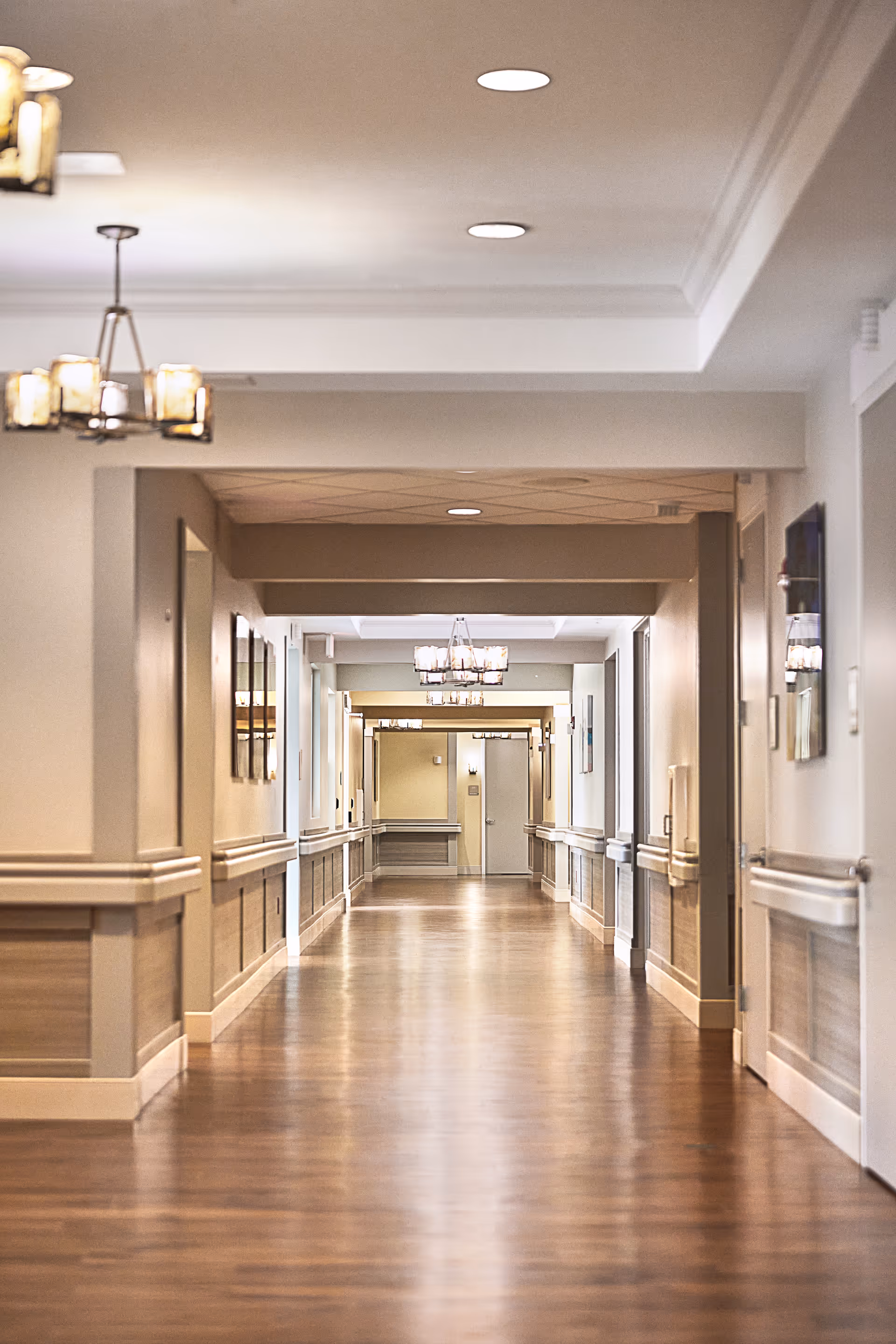 Long bright hallway inside a senior living facility with wood flooring and chandeliers.