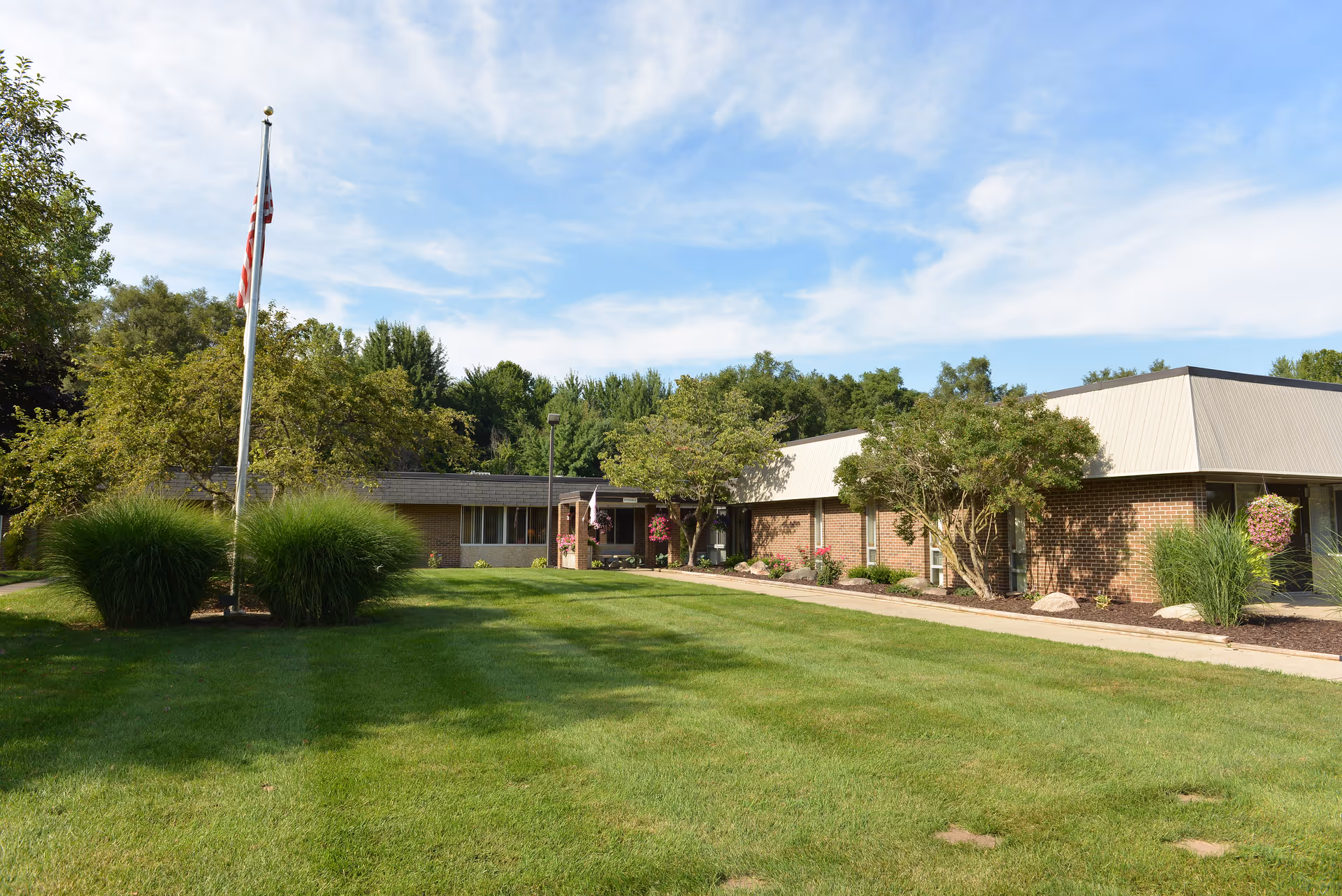Brick senior living facility with neatly manicured lawn, flagpole, and trees along the front entrance.
