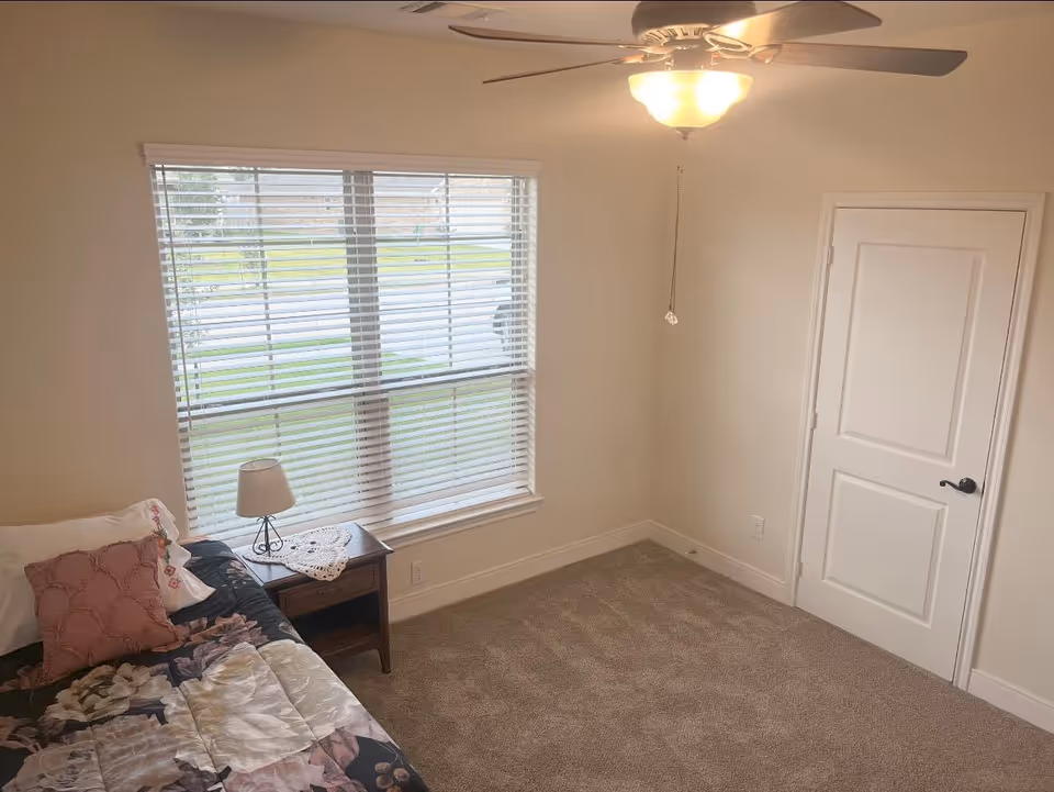Sunlit bedroom with bed, nightstand, lamp, window blinds, ceiling fan, and door.