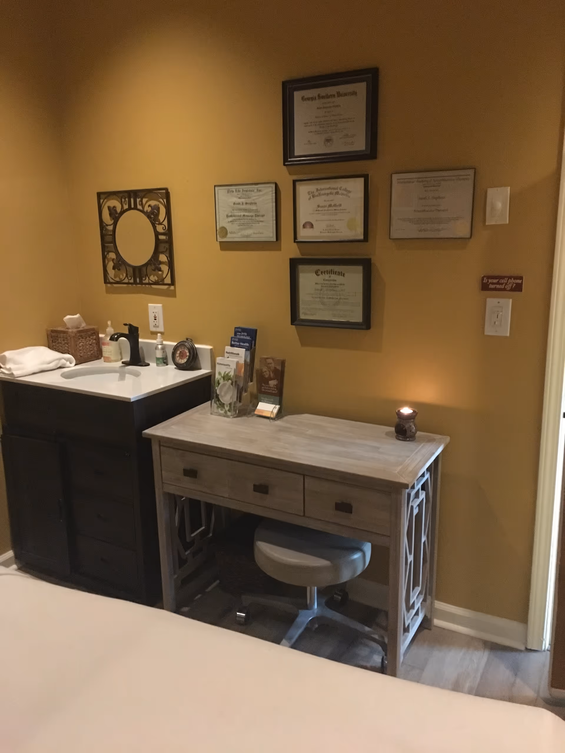 Massage therapy room with wooden desk, sink, and framed certificates on tan walls.