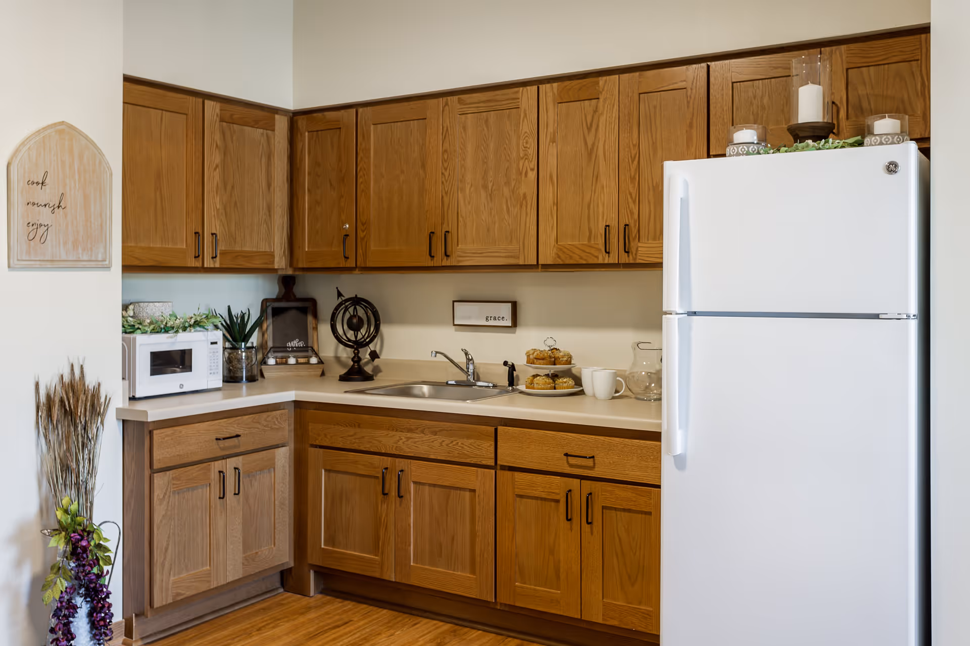 A kitchen area with wooden cabinets, a white refrigerator, a microwave, a sink, and various decorative items including candles, a small globe, and a plate of muffins. There is a wall decoration with the words 'cook, nourish, enjoy' and a small sign above the sink that says 'grace.'