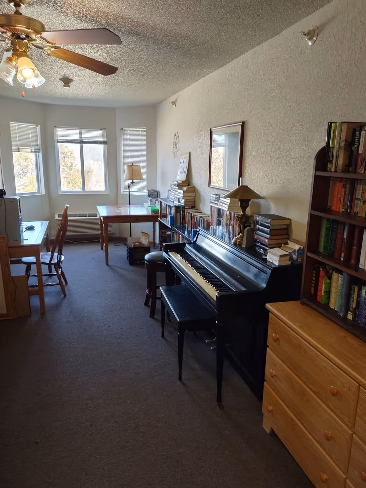 A long living room with an upright piano, bookshelves, a desk and chairs beneath bay windows and a ceiling fan.