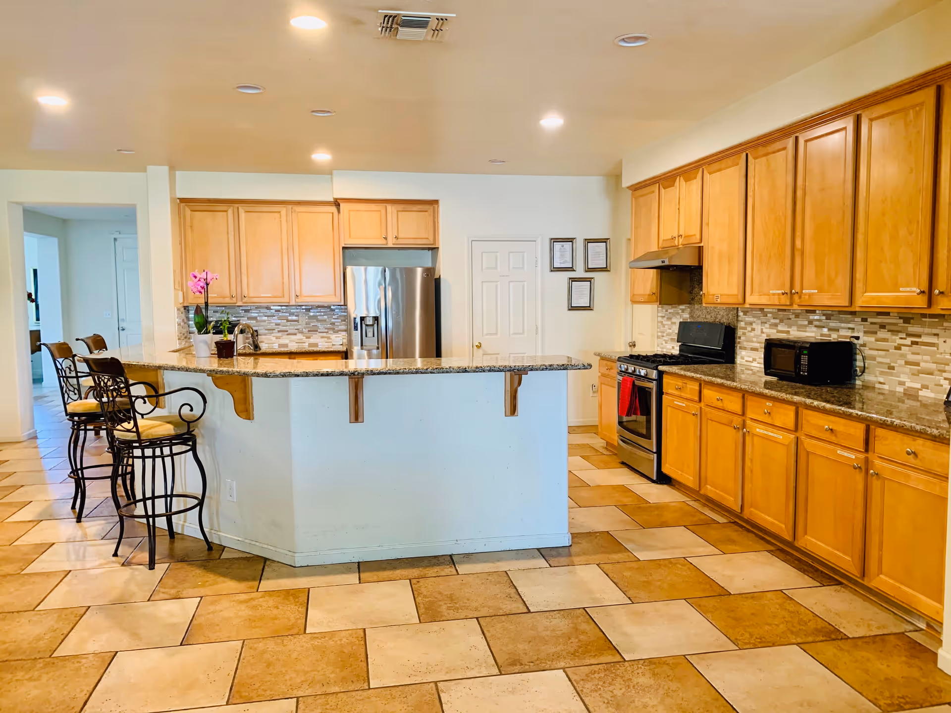 A spacious kitchen with light wooden cabinets, a granite countertop island with three wrought iron bar stools, stainless steel refrigerator and stove, a microwave on the counter, and tiled floor in beige and brown tones. The kitchen is well-lit with recessed ceiling lights.