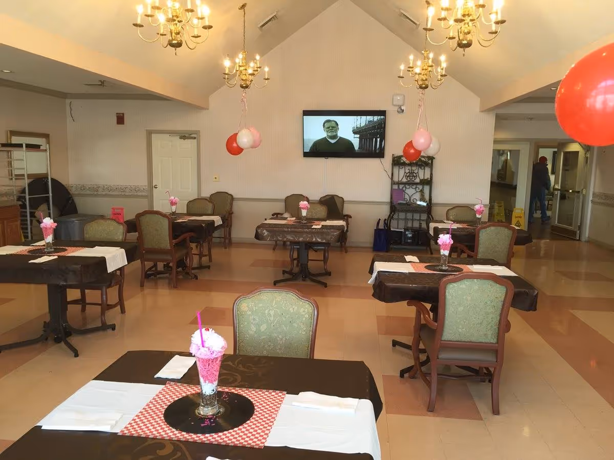 A dining room with multiple tables covered in dark tablecloths and white runners, each set with napkins and a glass with pink decorations and a straw. The room has green upholstered chairs, chandeliers hanging from the ceiling, red and white balloons tied to some chairs, and a television mounted on the far wall displaying a man. There is a shelving unit against the wall and a person visible in the hallway on the right side.