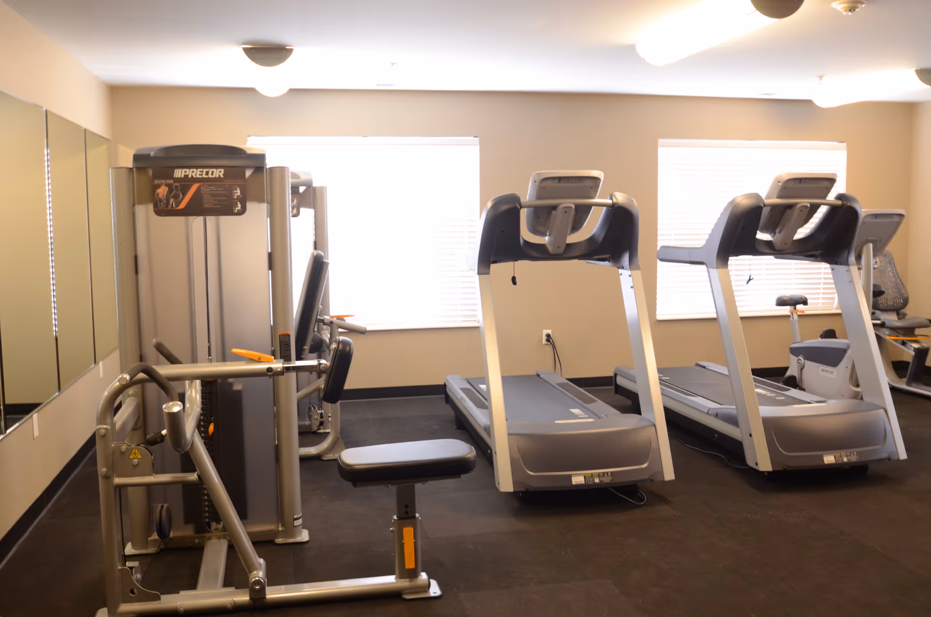 A fitness room with exercise equipment including two treadmills, a weight machine, and a stationary bike. The room has beige walls, two windows with blinds, and mirrors on one wall.