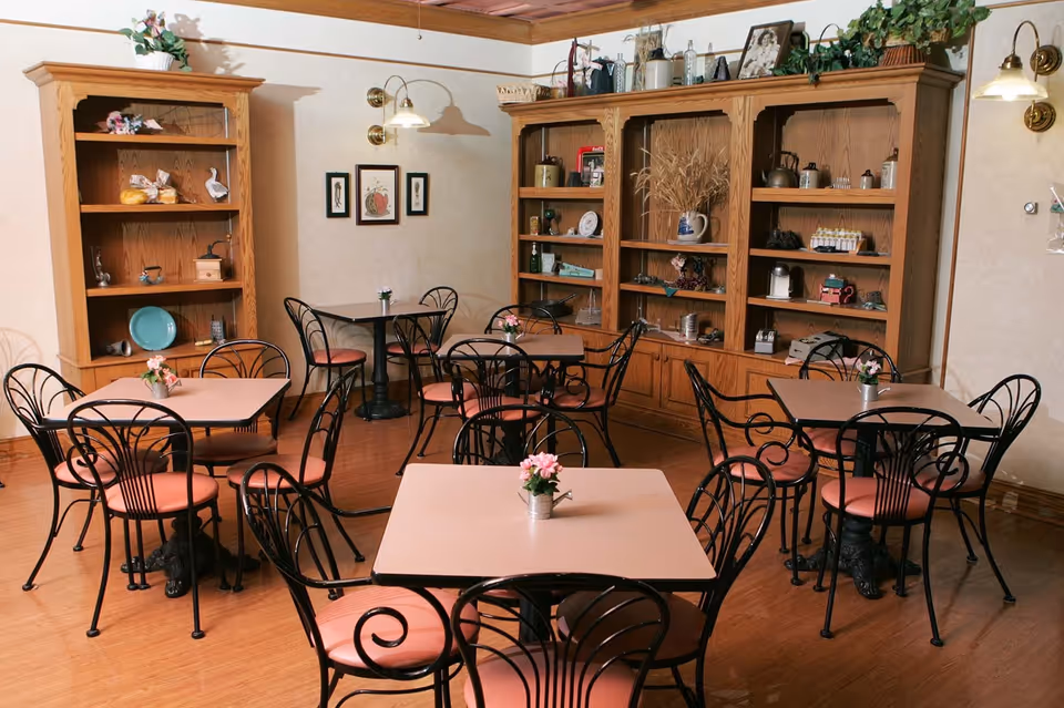 A cozy dining room with several square tables, each surrounded by black metal chairs with pink cushions. The room features wooden flooring, built-in wooden shelves filled with decorative items, and wall-mounted lamps providing warm lighting. Small flower arrangements are placed on each table.