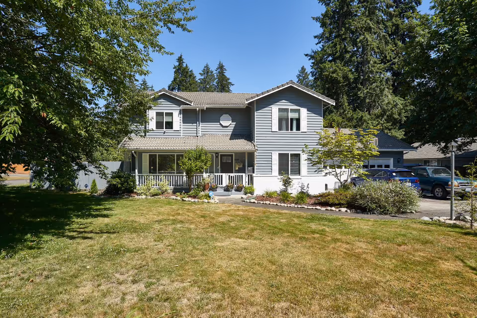 A two-story gray house with white trim surrounded by a well-maintained lawn and various trees and shrubs under a clear blue sky. There are several cars parked on the driveway to the right of the house.