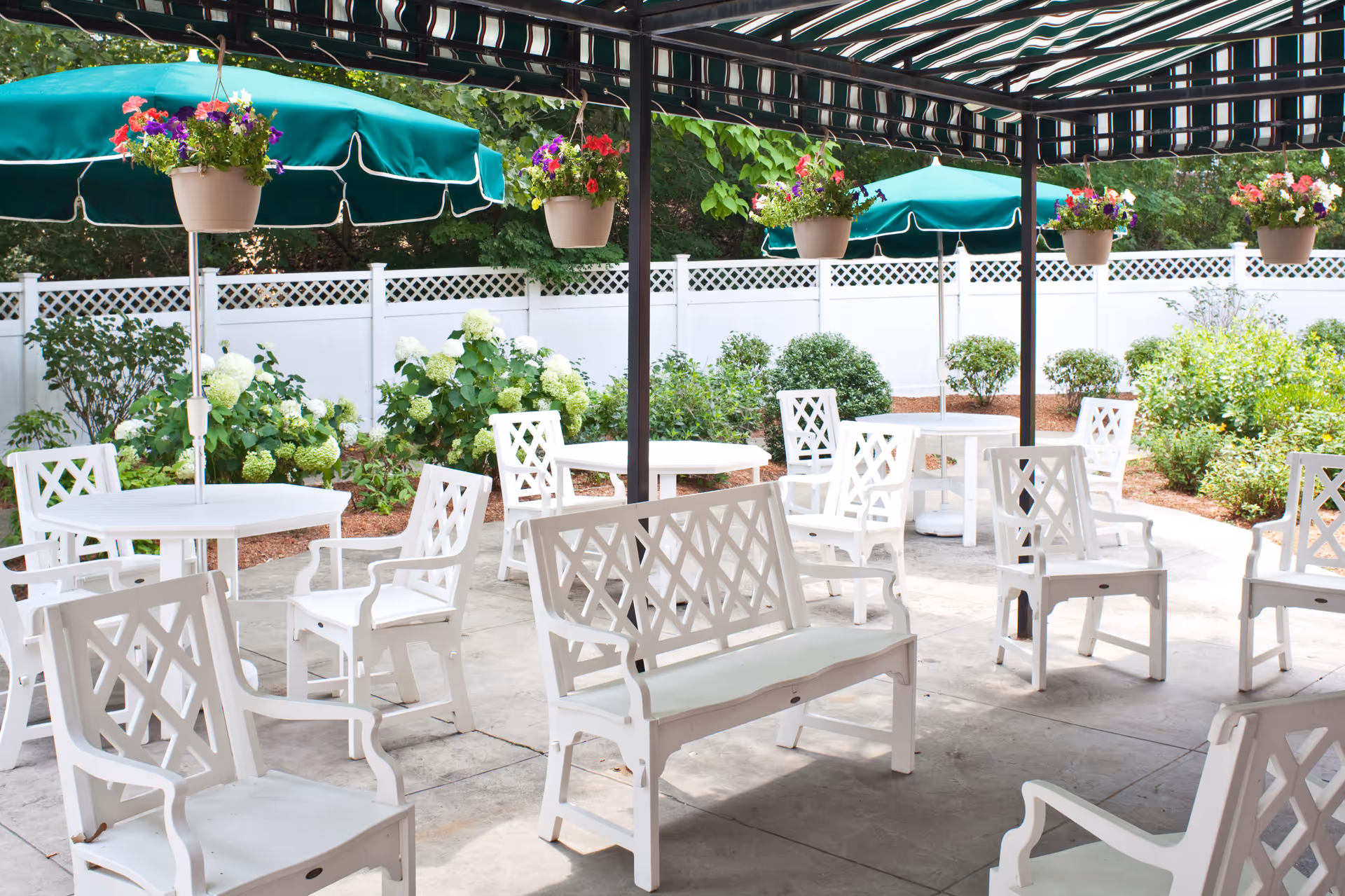 Outdoor patio area with white chairs and tables, some shaded by green umbrellas. Hanging flower pots with colorful flowers are suspended from a pergola. The area is surrounded by a white fence and lush green plants and bushes.