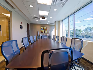 A long wooden table surrounded by blue chairs in a bright, windowed conference room.