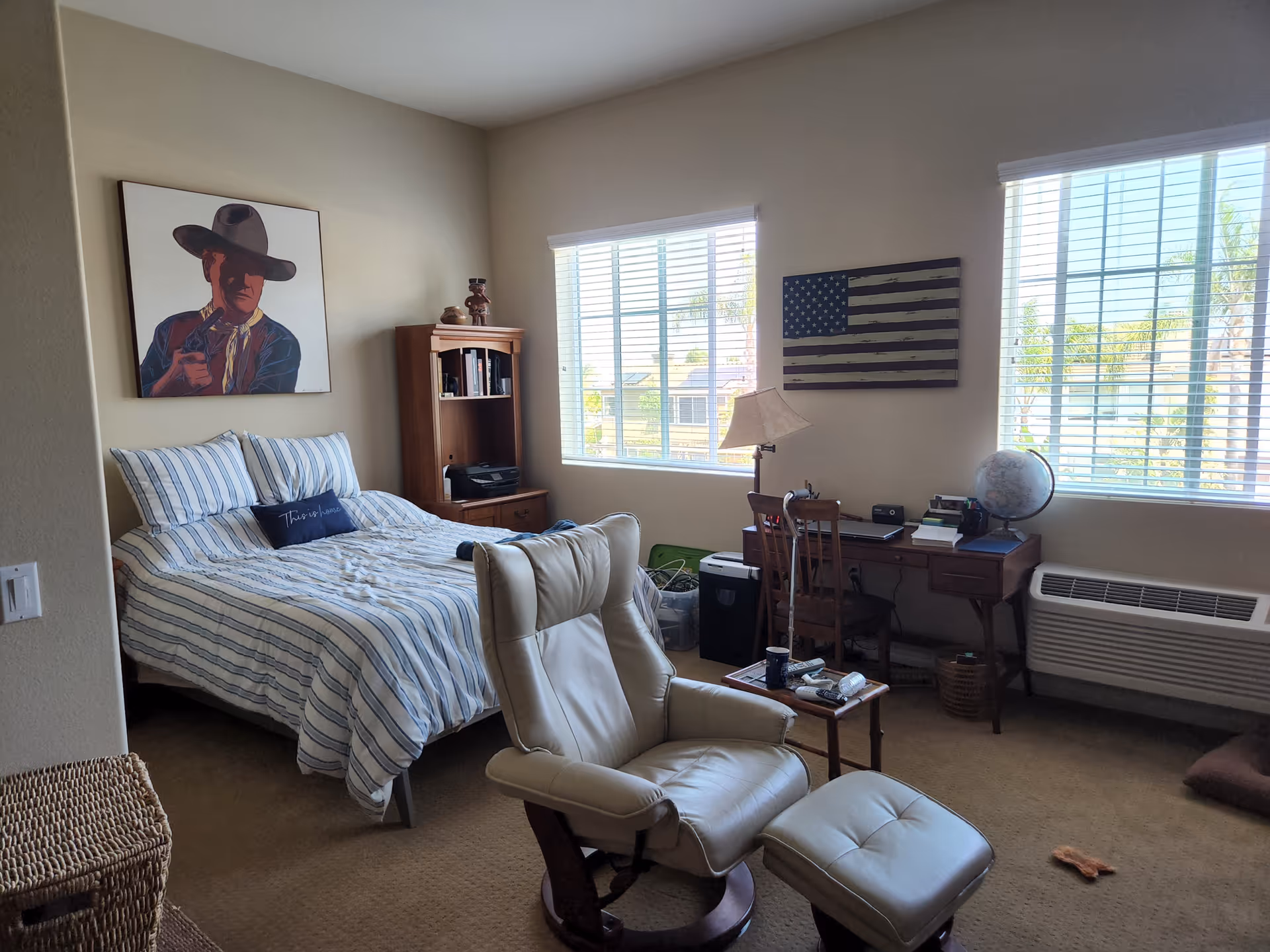 Bedroom in a senior living unit with striped bed, recliner, desk, globe, and American flag art.
