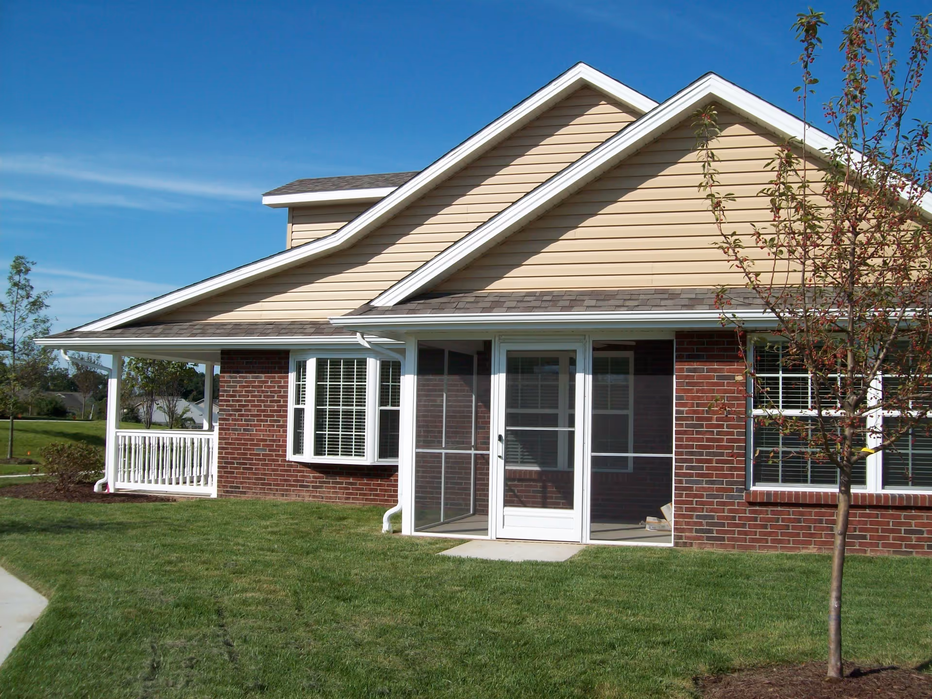 Front exterior of a single-story brick and siding senior living unit with a screened porch, windows, and a small lawn.