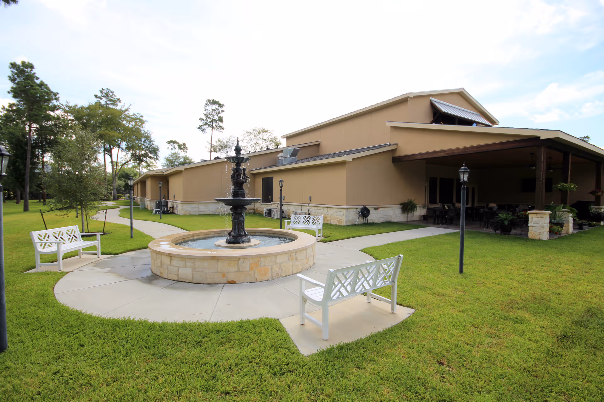 Outdoor senior living courtyard with central fountain, white benches, and a beige building entrance.
