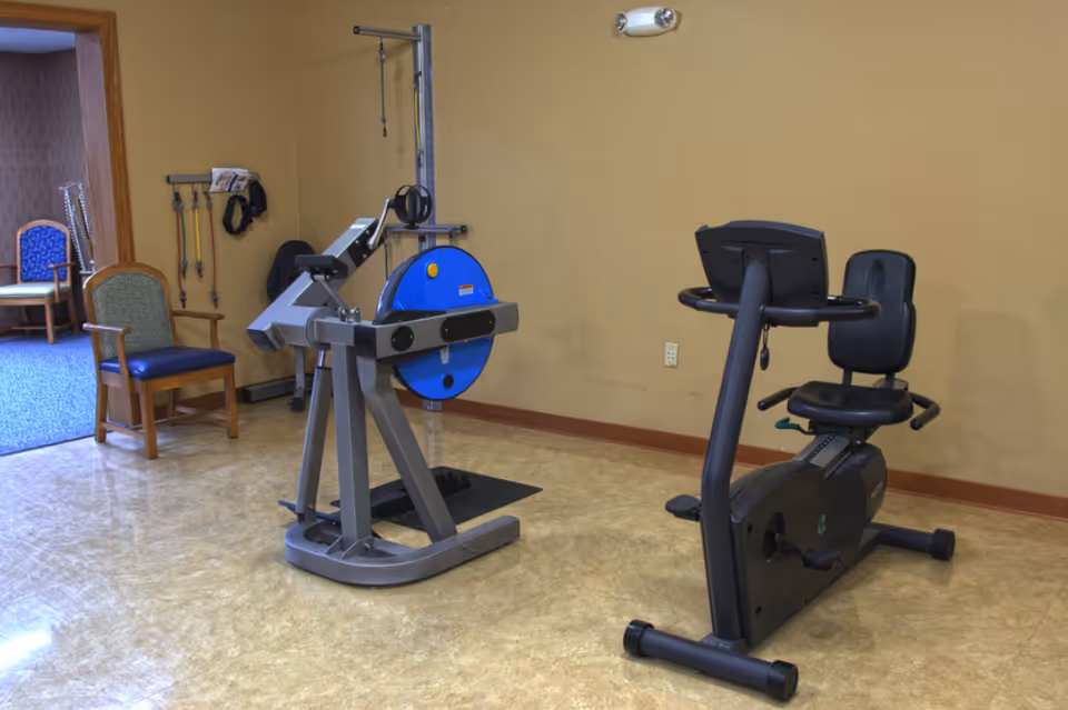 A small exercise room with two pieces of fitness equipment, including a recumbent stationary bike and a resistance training machine. There are two chairs against the wall, and various exercise bands and straps hanging on the wall in the background. The room has beige walls and a light-colored floor.