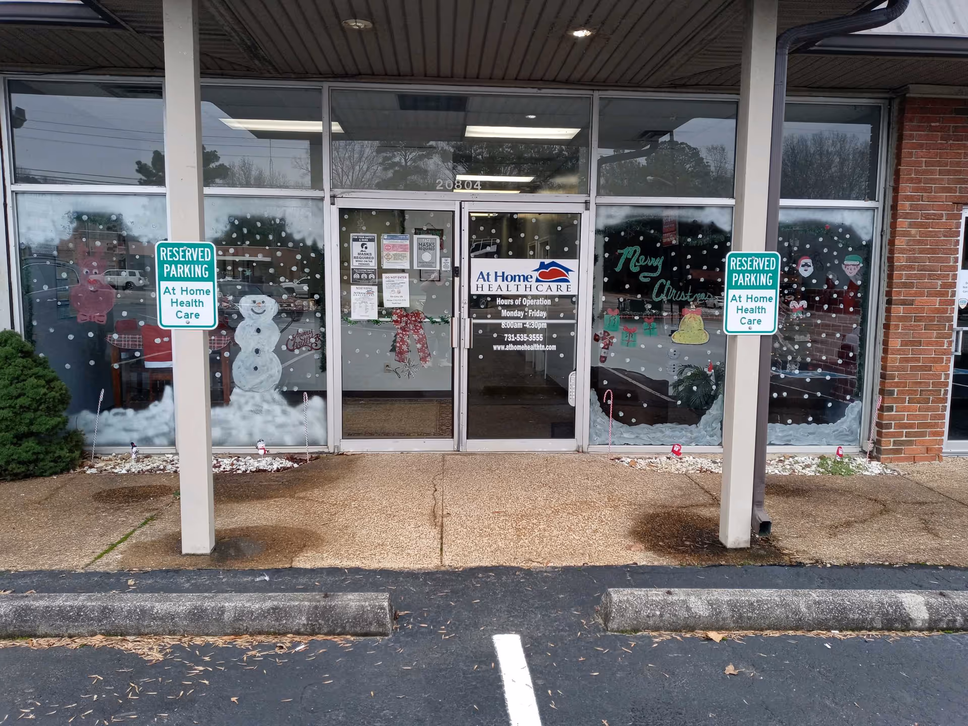 Storefront entrance of At Home Healthcare with holiday window decals and reserved parking signs.