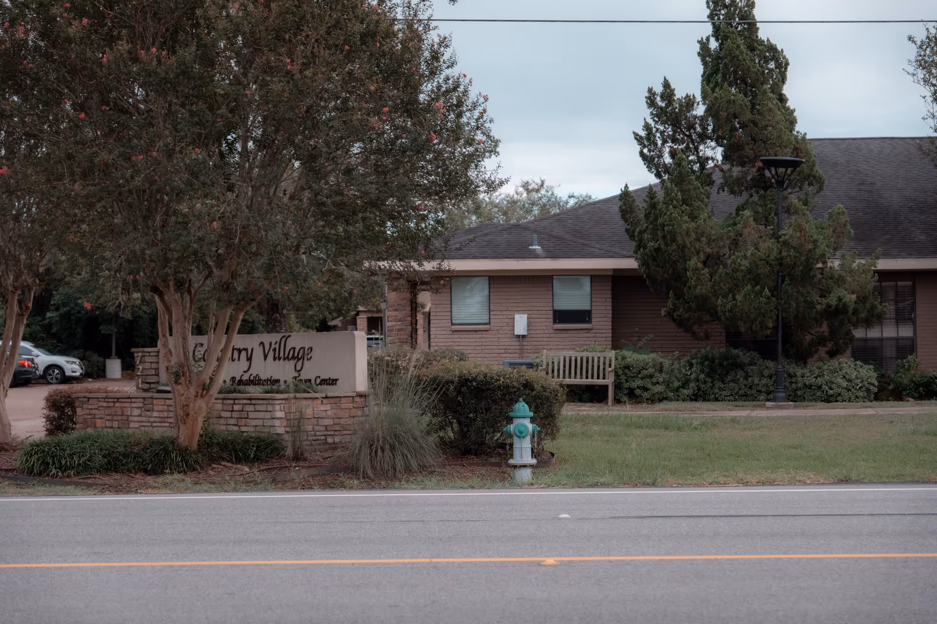 Front exterior of the Country Village building showing the entrance sign, landscaping, bench, and a green fire hydrant by the road.