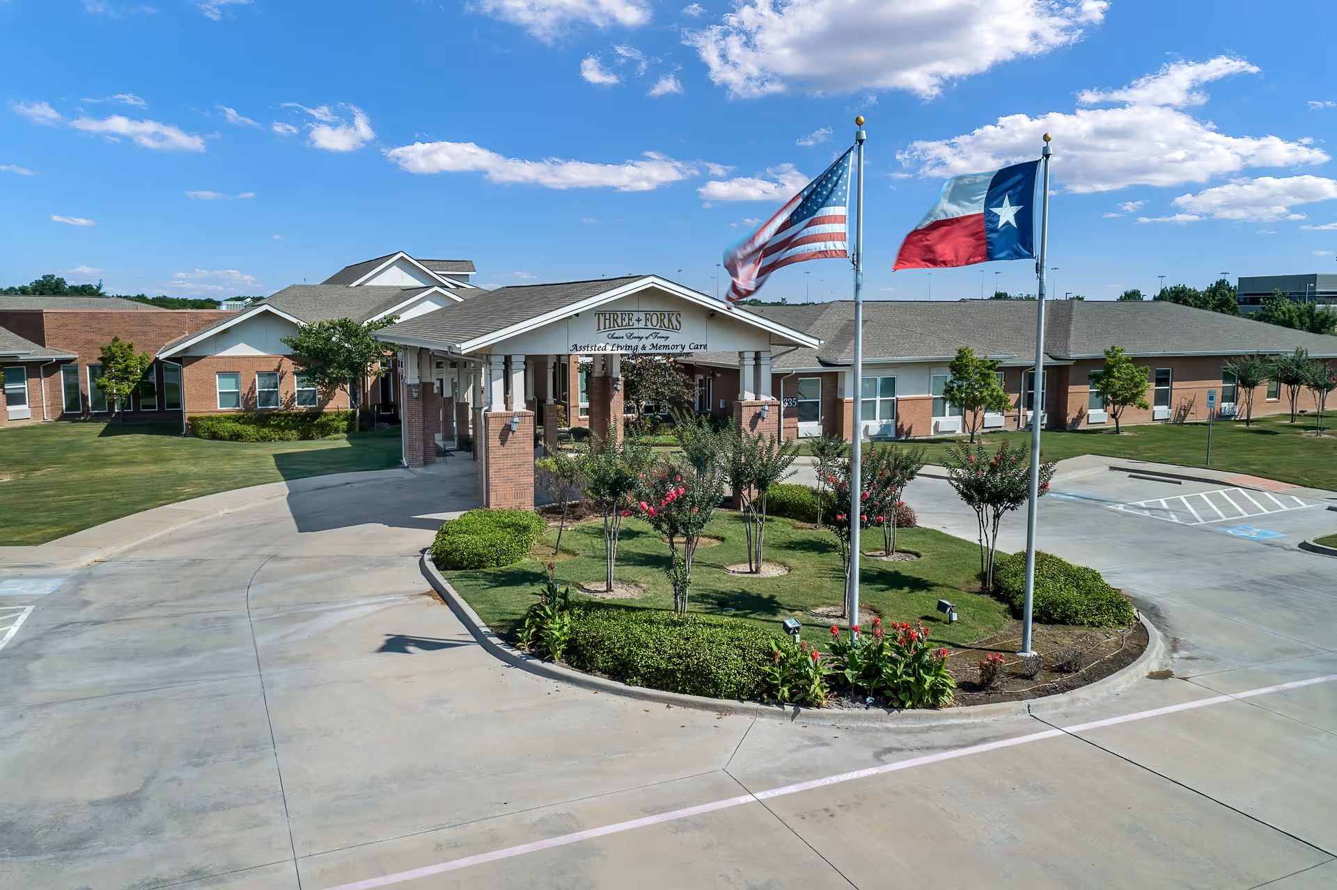 Front entrance of a single-story senior living facility with a covered porte-cochère, three flagpoles, and a landscaped circular driveway.