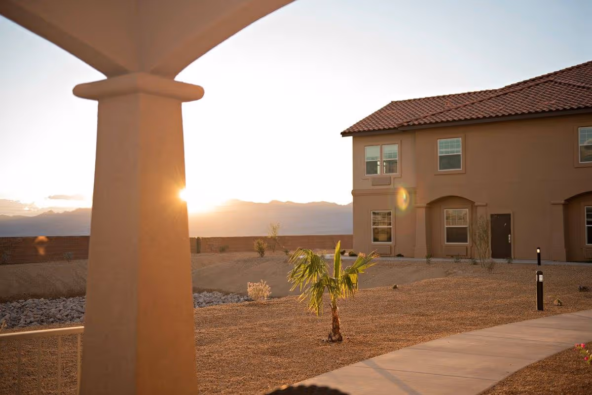 Sunlit exterior view of Joshua Springs Senior Living, showing a stucco building, walkway, and desert landscaping.