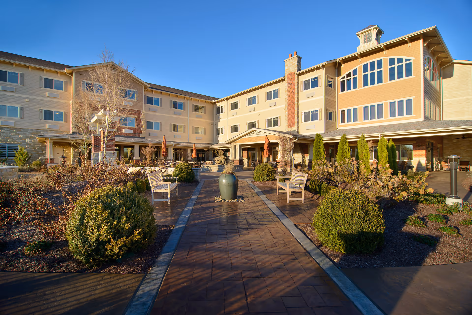 Exterior view of The Bridge at Hickory Woods senior living facility showing a multi-story building with numerous windows, a paved walkway lined with benches and greenery, and outdoor seating with umbrellas under a clear blue sky.