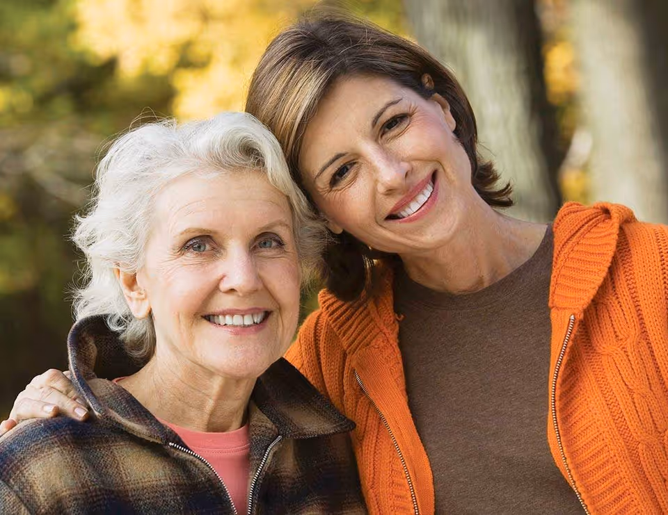 Two smiling women pose together outdoors with autumn trees in background