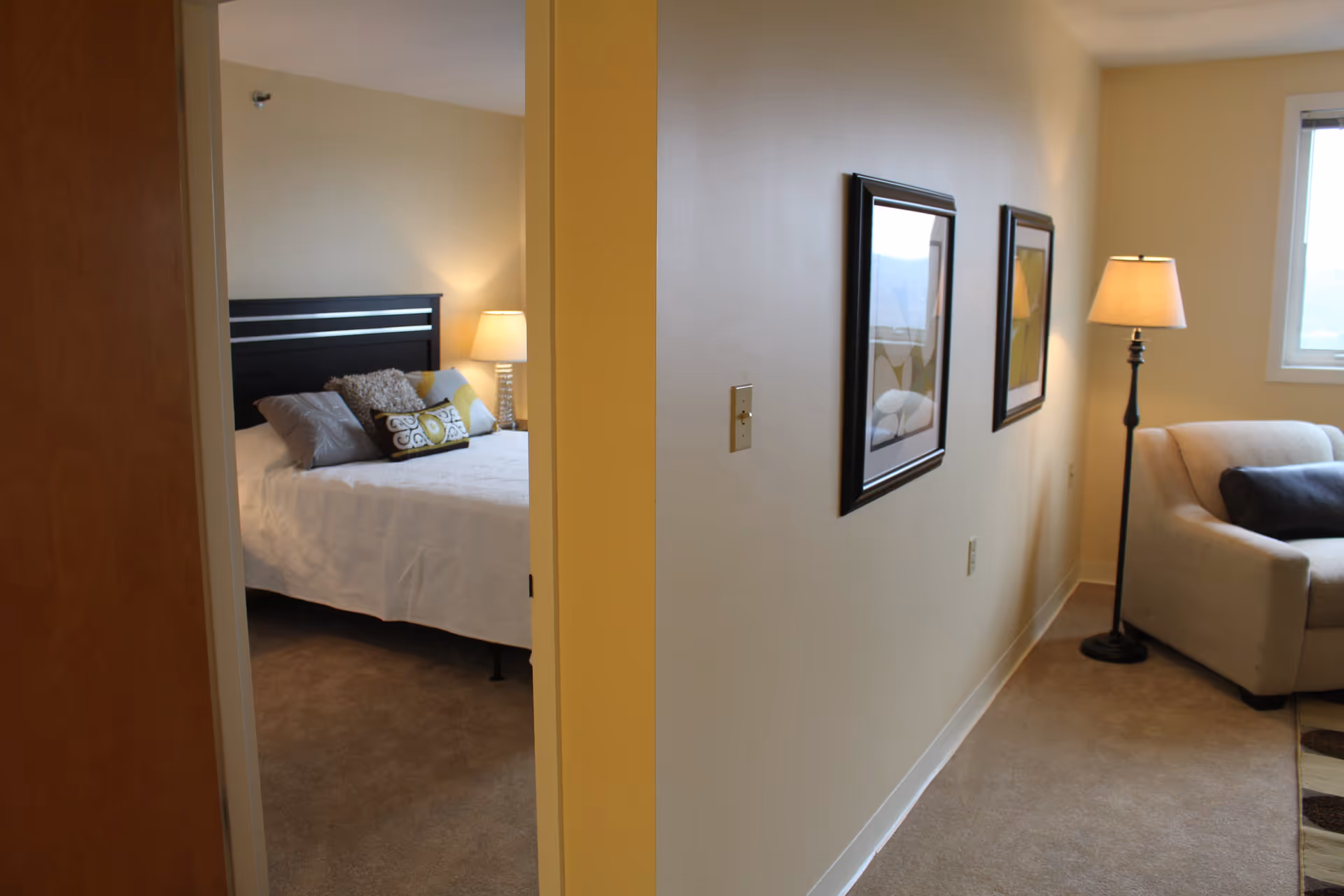 View of a cozy interior space showing a bedroom on the left with a bed adorned with multiple pillows and a bedside lamp, and a living area on the right with a beige armchair, a floor lamp, two framed pictures on the wall, and a window letting in natural light.