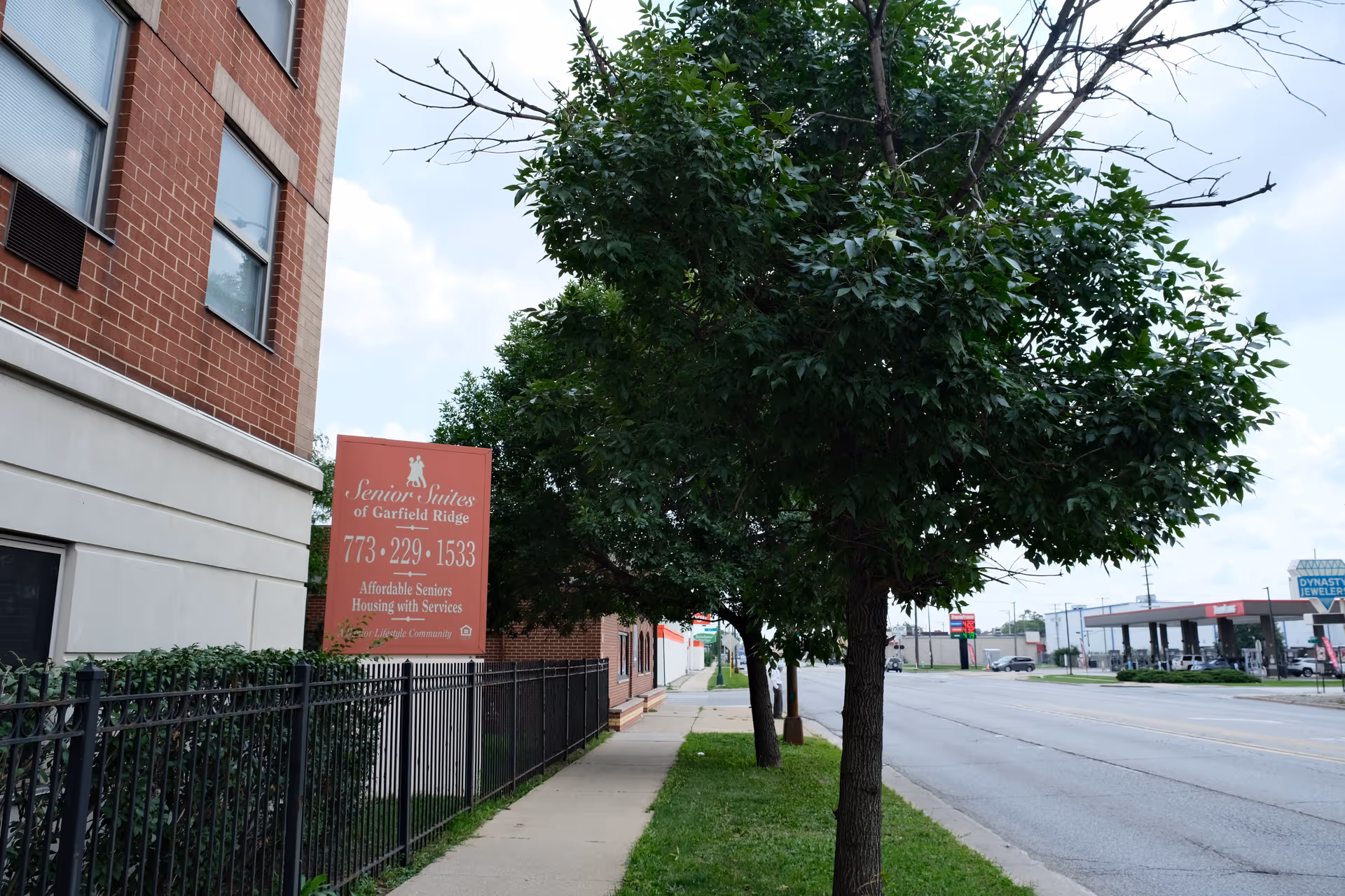 Street view of the Senior Suites of Garfield Ridge building with its red sign, sidewalk, and trees along the road.