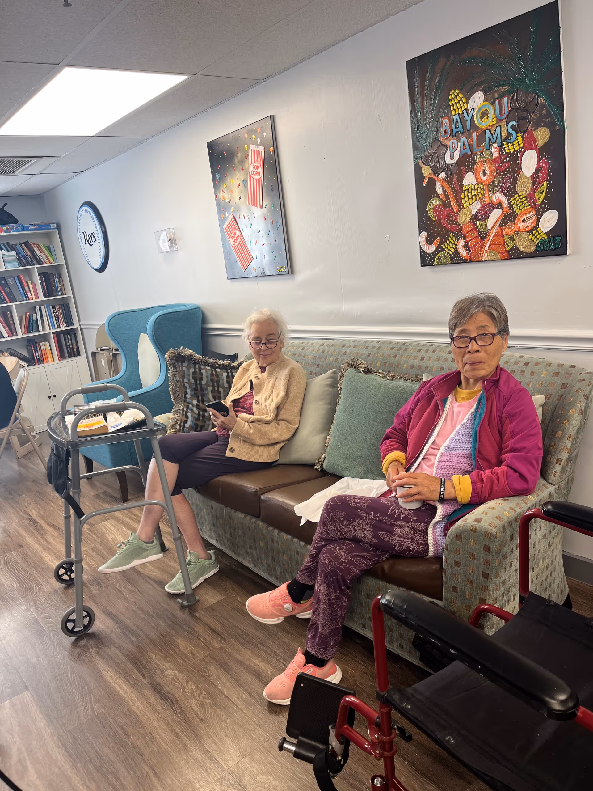 Two elderly women sit on a patterned sofa in a bright assisted living lounge.