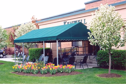 Outdoor patio area with green canopy, chairs, and blooming flower bed in front of building.
