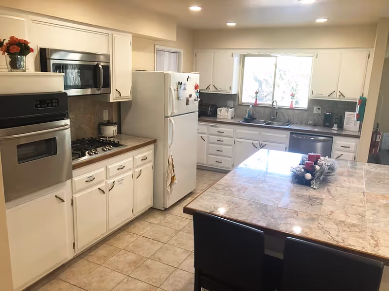A bright kitchen with white cabinets, a white refrigerator, a built-in oven, a microwave, a gas stove, and a dishwasher. There is a large tiled island countertop with two black chairs. The kitchen has a window above the sink letting in natural light and some decorative items on the counter and island.