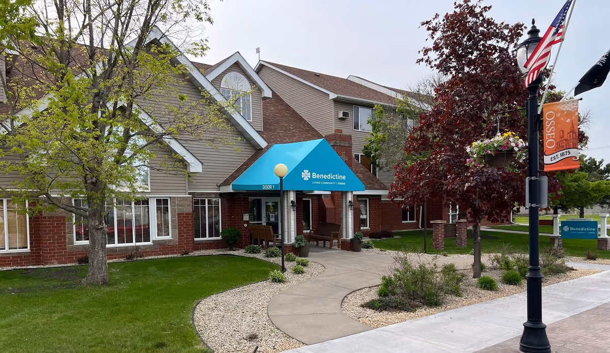 Exterior view of Benedictine Living Community-Osseo building entrance with a blue awning, surrounded by green grass, trees, and a curved sidewalk. An American flag and a banner with 'Osseo EST. 1875' are visible on a lamp post near the sidewalk.