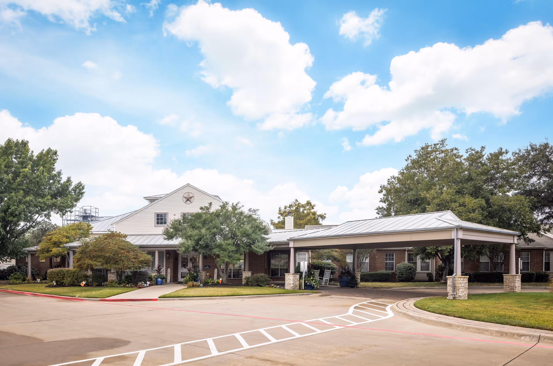 Front exterior of a single-story senior living facility with a covered drop-off porte-cochère, landscaped greenery, and a partly cloudy blue sky.