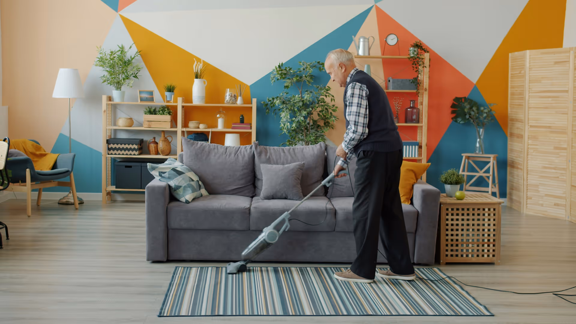 Elderly man vacuuming a colorful living room with sofa and plants.
