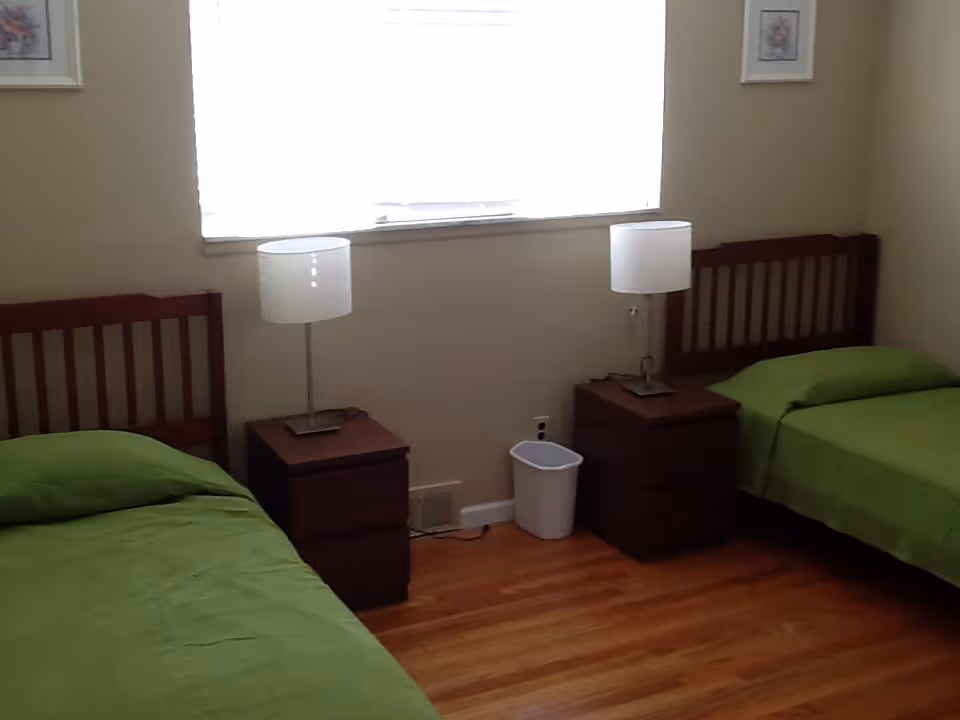 A simple bedroom with two single beds, each with a green bedspread. Between the beds are two wooden nightstands, each with a white lamp. There is a small white trash bin on the floor between the nightstands. The room has wooden flooring and a window with blinds letting in light. Two framed pictures hang on the walls above each bed.