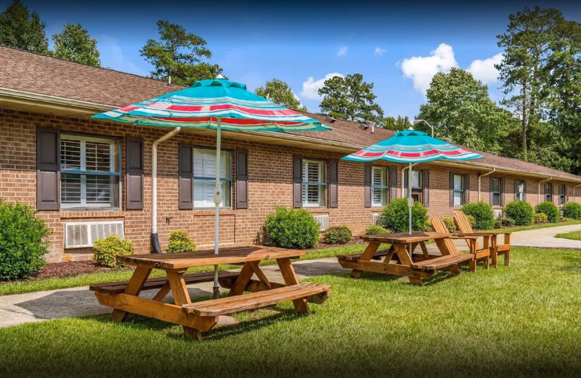 Brick senior living building with picnic tables and colorful umbrellas outdoors on green lawn.