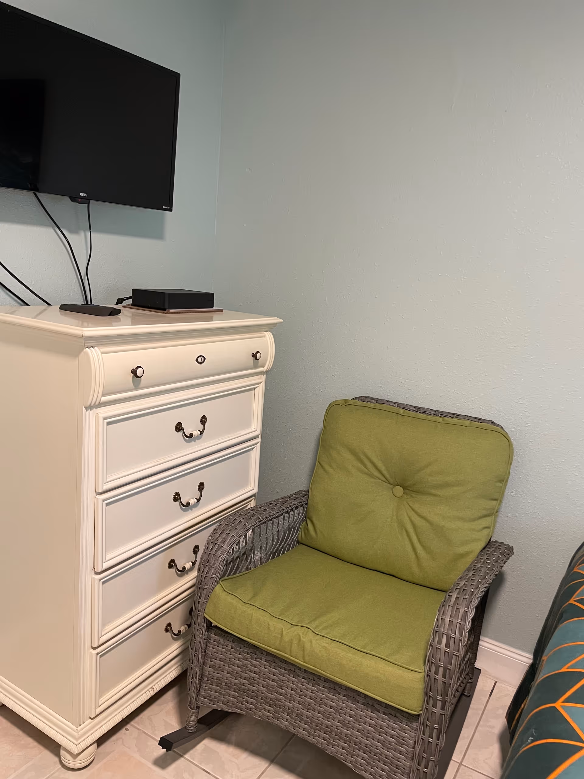 A white dresser with drawers sits beside a wicker chair with green cushion, mounted TV above.