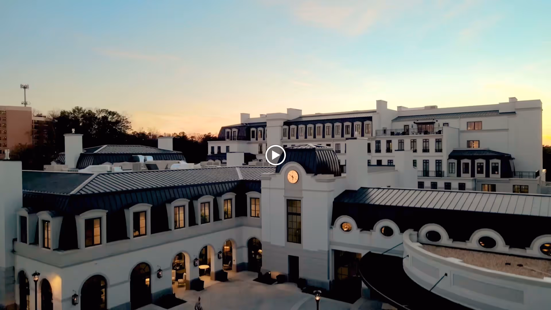 Exterior view of a large, elegant multi-story building at sunset with a clock tower in the center, featuring arched windows and a courtyard area with outdoor lighting.
