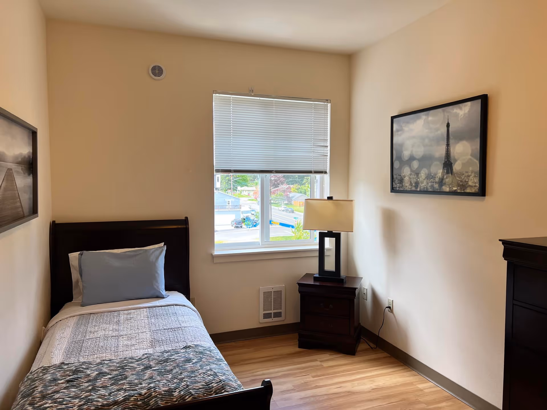 A small bedroom with a single bed, nightstand, lamp, window, and wall art.