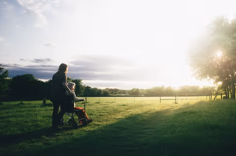 A caregiver pushes an elderly person in a wheelchair across a sunlit grassy field outdoors.