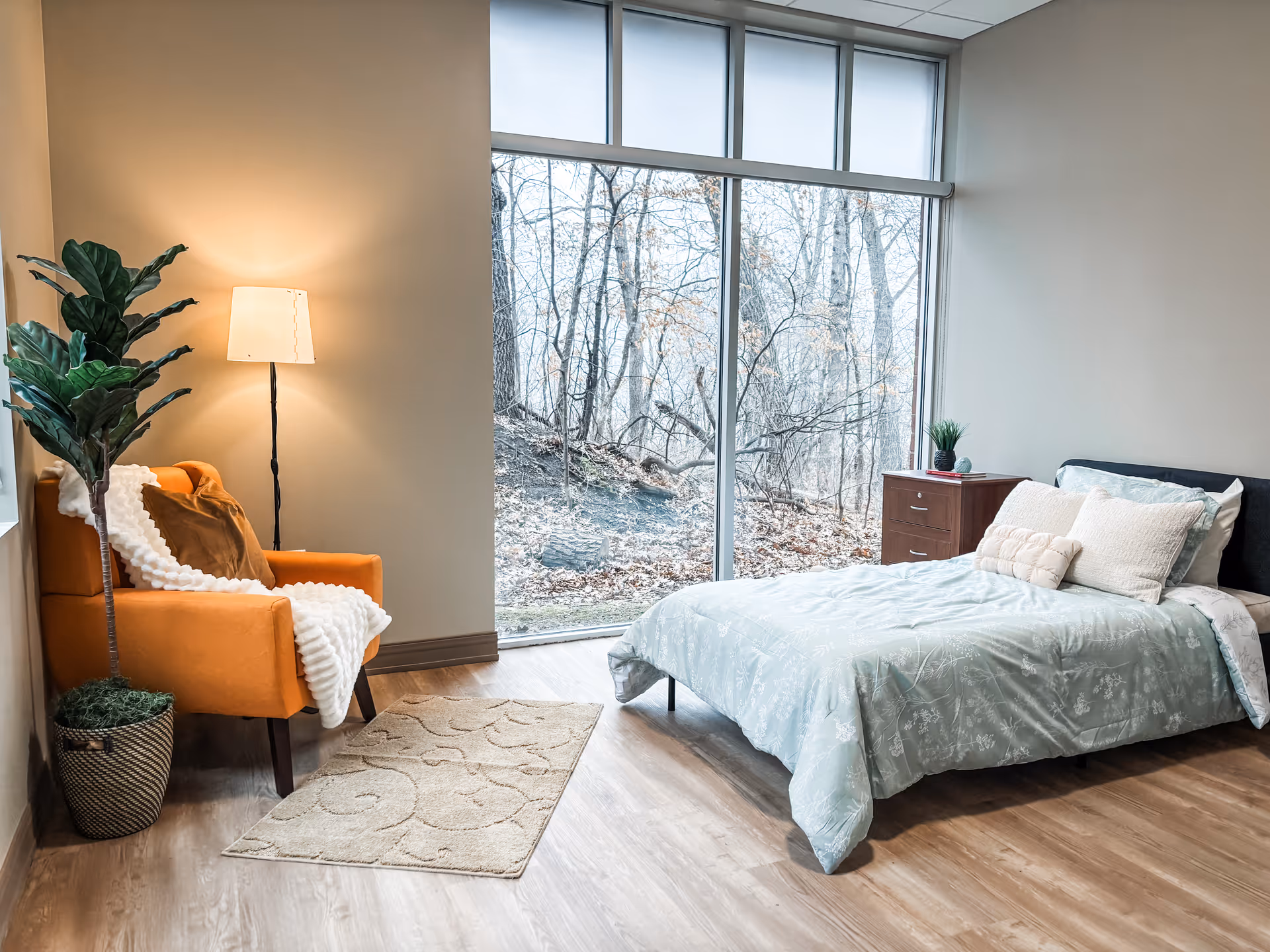 Bright bedroom with bed, orange chair, lamp, and large window overlooking trees.