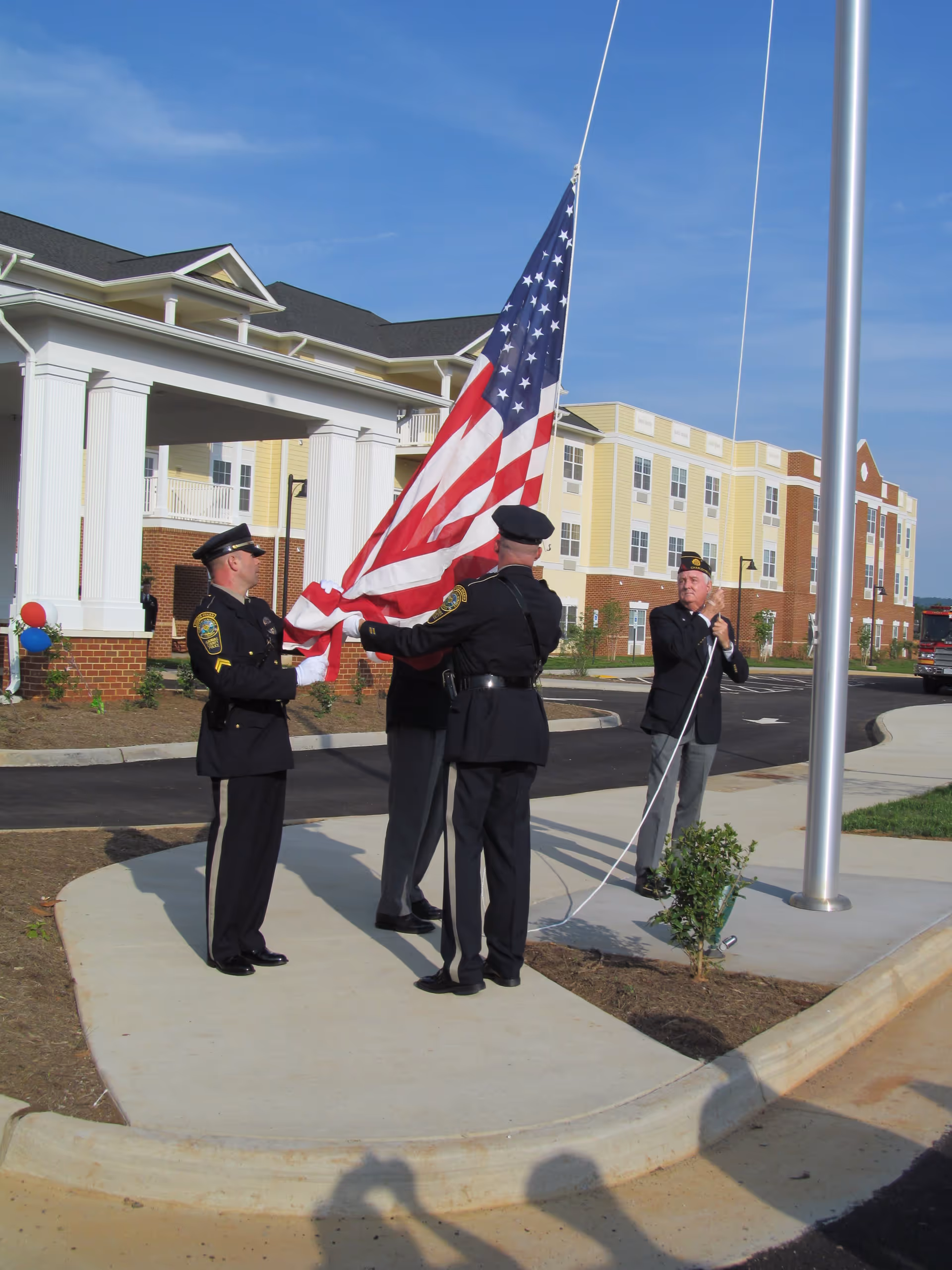 Uniformed officers raise an American flag at a flagpole in front of a senior living building.