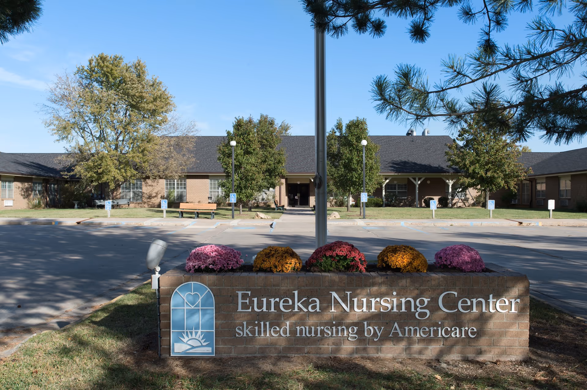 Exterior view of Eureka Nursing Center building with a brick sign in front that reads 'Eureka Nursing Center skilled nursing by Americare'. The sign is decorated with colorful flowers and there are trees and a parking area in the background under a clear blue sky.