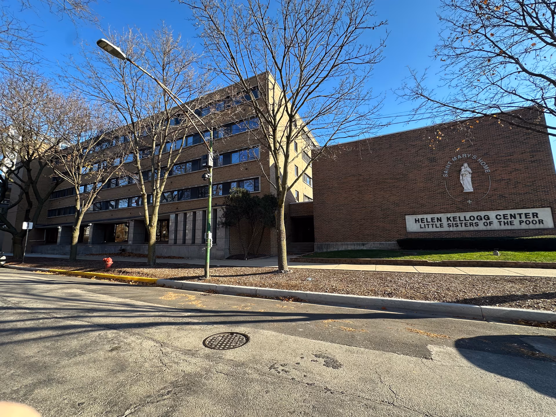 Exterior view of a multi-story brick building with a sign that reads 'Helen Kellogg Center Little Sisters of the Poor' and a circular emblem above it that says 'Saint Mary's Home'. Leafless trees and a streetlamp are visible in front of the building under a clear blue sky.