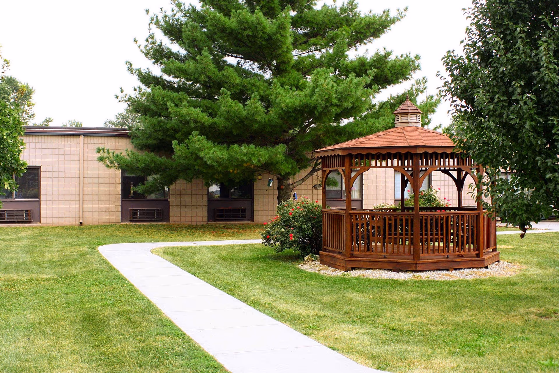 Outdoor area of a senior living facility with a wooden gazebo surrounded by green grass, trees, and a paved walkway leading towards a beige building with several windows.